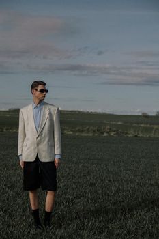 Man in sunglasses and blazer stands in a green field, offering a stylish contrast in a natural setting.