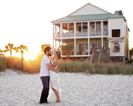 A couple embraces and dances joyfully on a sandy beach with a scenic beachfront house in the background during sunset.