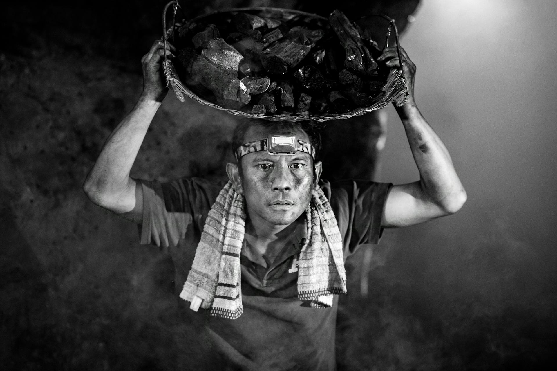 A black and white photo of a coal miner carrying a basket of coal on his head.