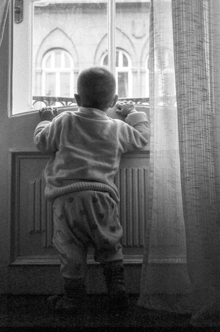 Black And White Photo Of A Baby Boy Standing By The Window In The Apartment