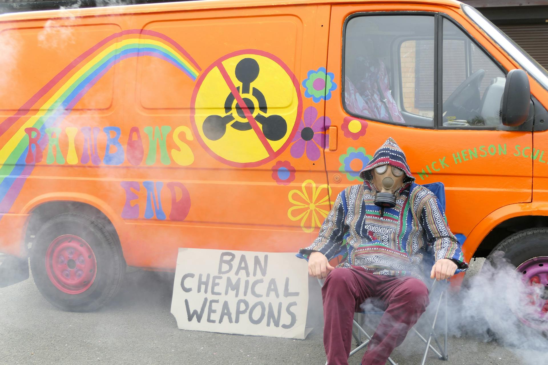 A protester in a gas mask sits by a colorfully painted van with an anti-chemical weapons sign.