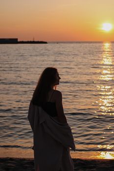 A woman wrapped in a blanket gazes at the ocean during a peaceful sunset.
