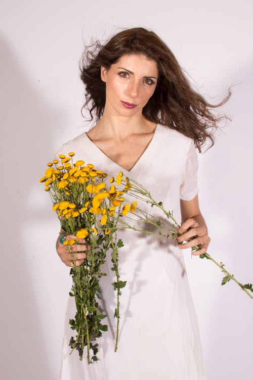 Woman in White Dress Holding Bunch of Yellow Flowers while Looking at the Camera