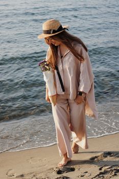 A woman in casual attire and a hat walks barefoot on a beach holding flowers, enjoying leisure by the sea.