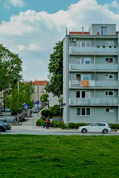 City street scene with apartment building, cars, and green space under a blue sky.
