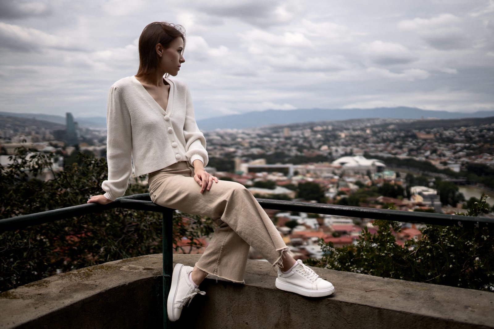 Woman in a cardigan and wide trousers sitting on a wall above a city