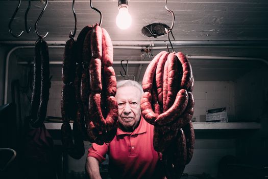 Elderly man in a butcher shop wearing a red shirt with hanging sausages in Mexico City.