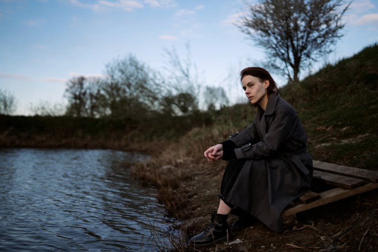 Woman In Coat Sitting On Lake Bank