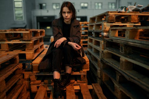 A Woman in Brown Coat Sitting on the Stack of Wooden Pallet while Looking at the Camera