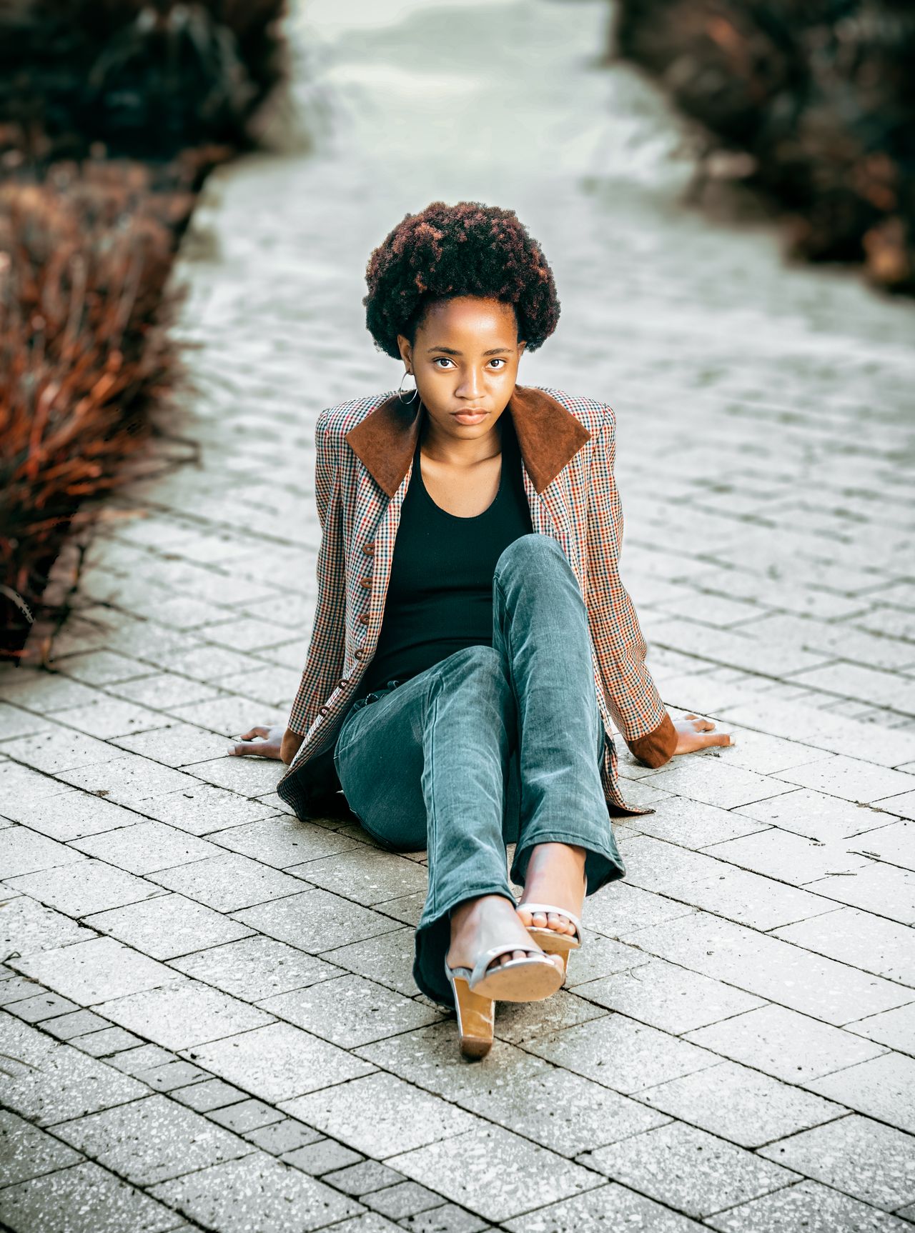 Woman in a checkered tweed blazer and jeans sitting on a paved path