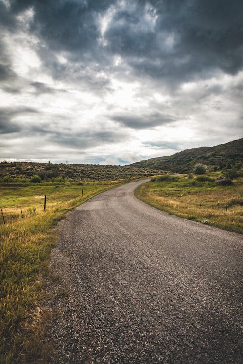 Free Gray Concrete Road Between Green Grass Field Under Gray Cloudy Sky Stock Photo