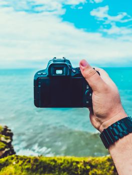 A hand holding a DSLR camera with an ocean view background in Torres, Brazil.