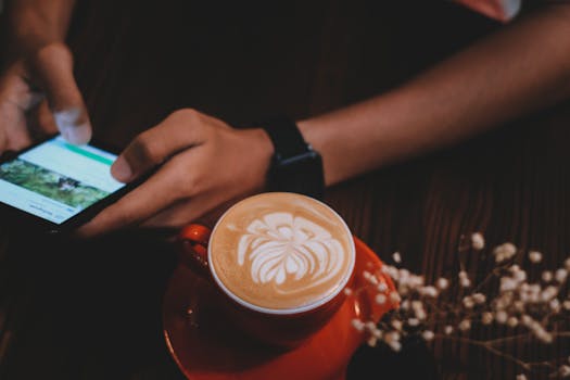High-angle view of a latte with beautiful art next to a person using a smartphone in a cozy cafe.