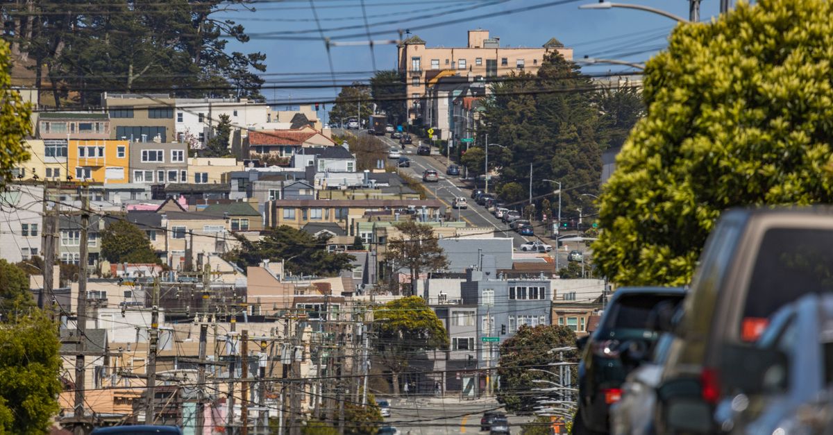 View of a city street filled with parked cars and residential homes, under a clear blue sky.