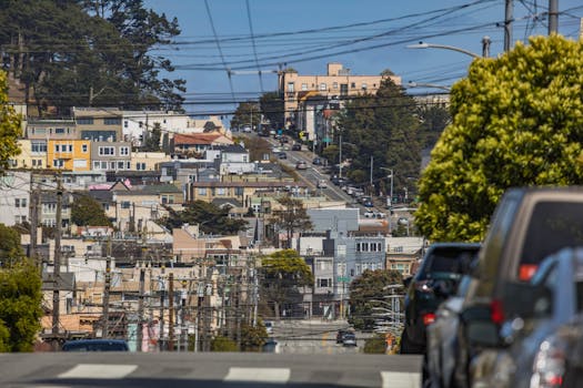 View of a city street filled with parked cars and residential homes, under a clear blue sky.