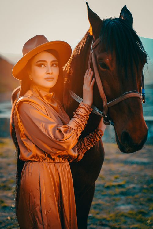 Free A woman in a stylish brown dress poses with a horse during a sunset on a farm. Stock Photo