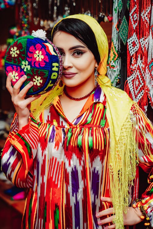 Free A Woman Wearing Yellow Scarf on Her Head Holding a Colorful Round Bag while Looking at the Camera Stock Photo