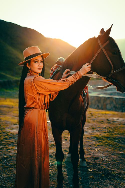 Free A woman in an orange dress and fedora stands with a horse at sunset. Stock Photo