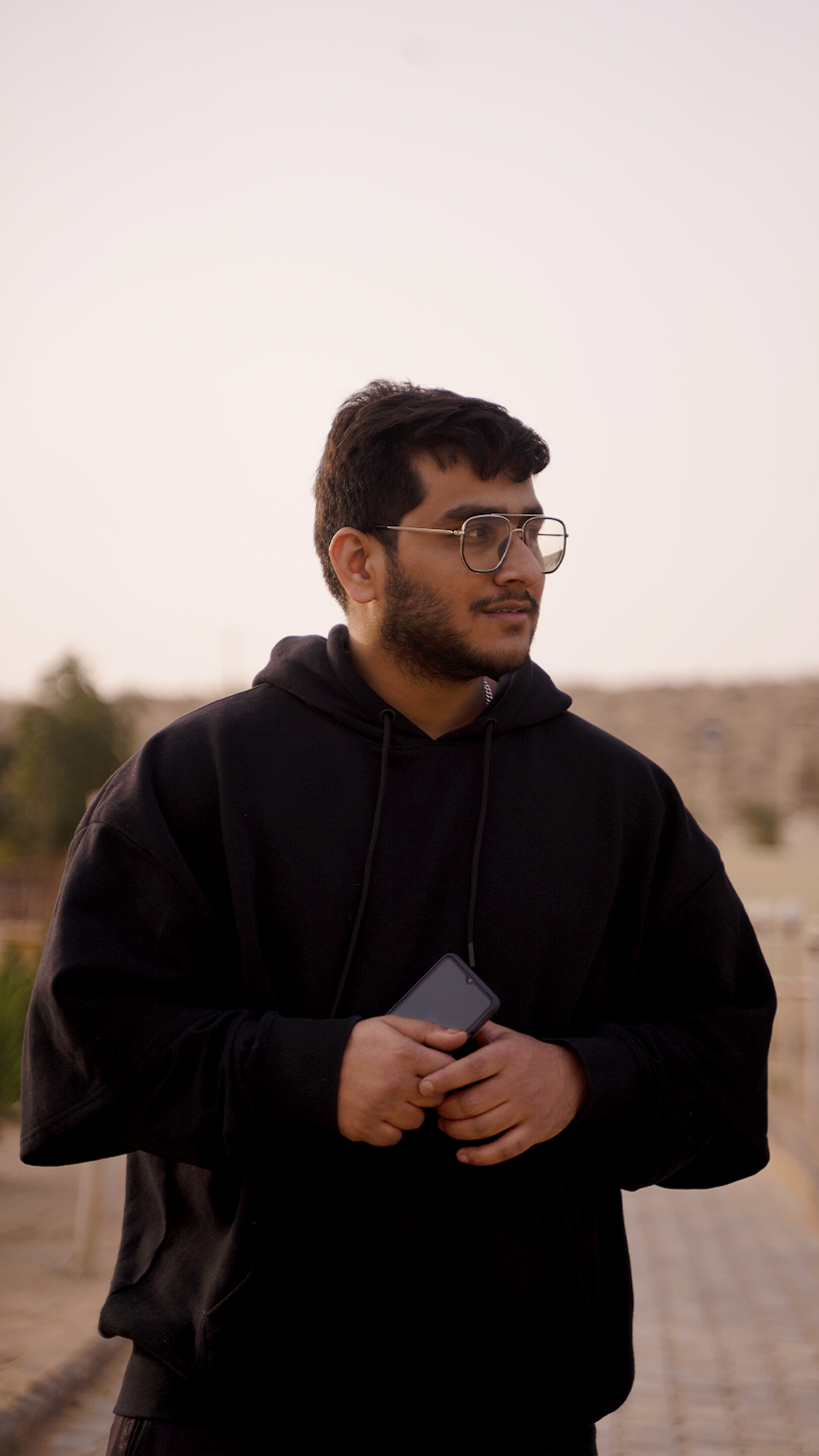 A bearded man in a hoodie holding a smartphone while looking into the distance outdoors.