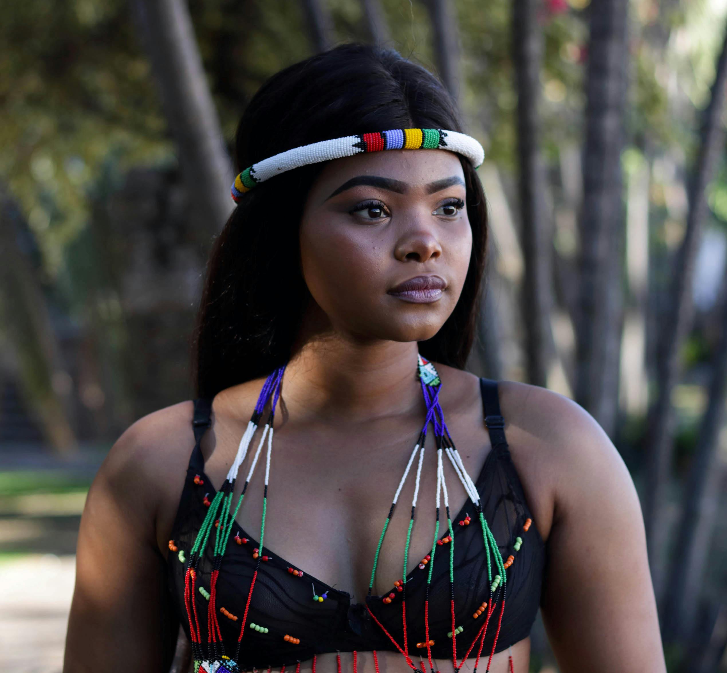 A young woman wearing colorful traditional beaded attire standing outdoors amid trees.