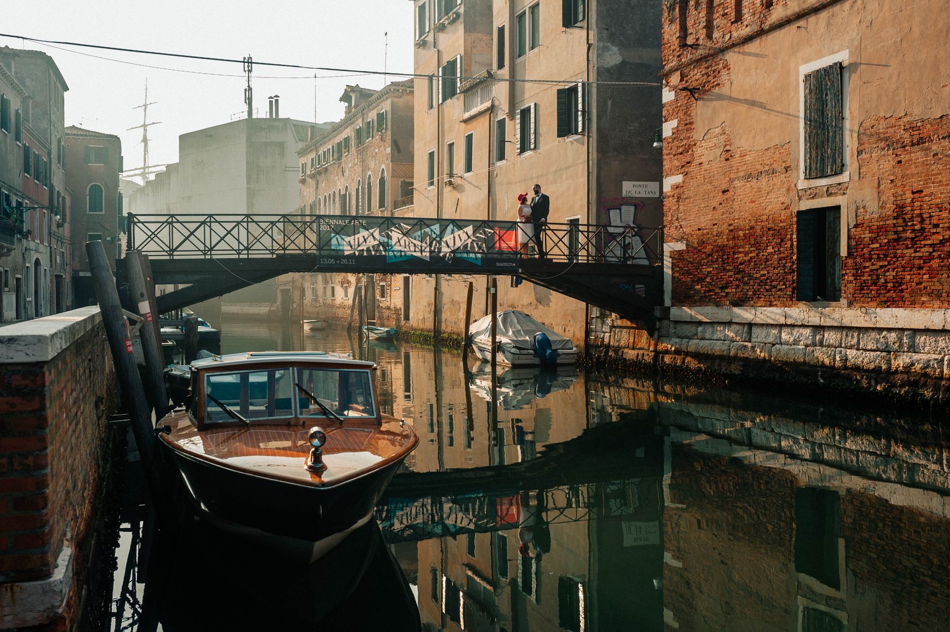 A romantic couple stands on a Venetian canal bridge at sunrise, with boats docked nearby.