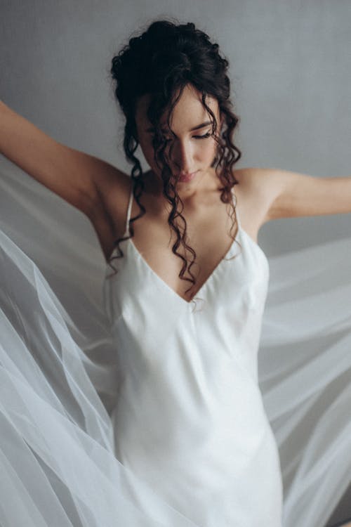 Woman in White Spaghetti Strap Dress Holding Veil while Looking Down