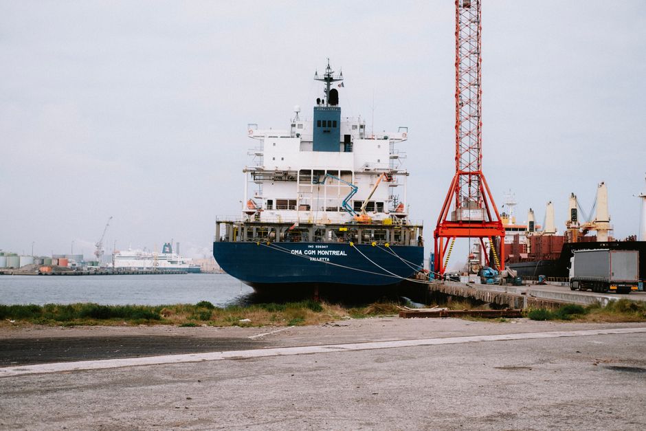 Large cargo ship docked at an industrial port with cranes and shipping containers.