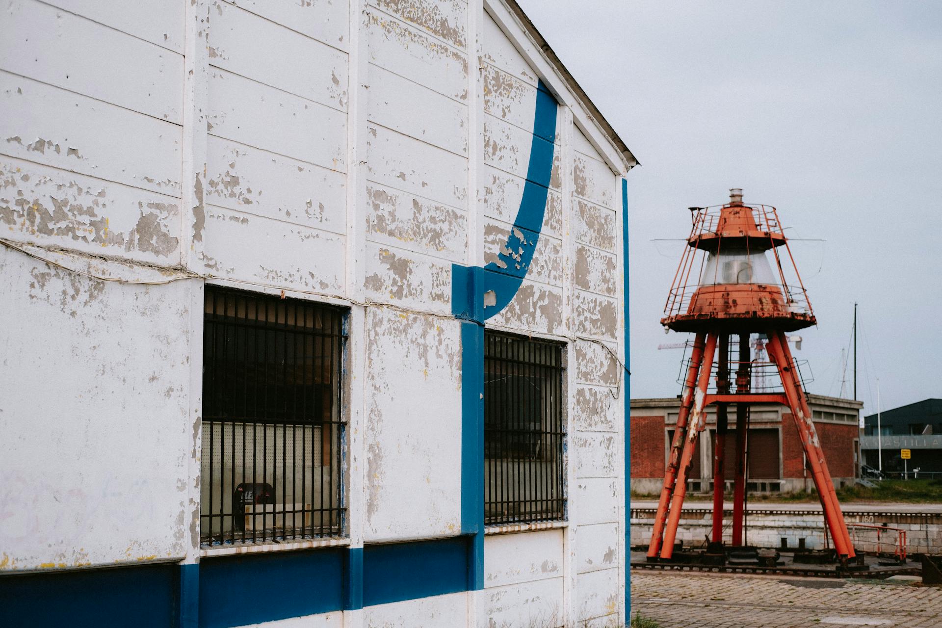 An old peeling building next to a rusty lighthouse against a gray sky, capturing decay.