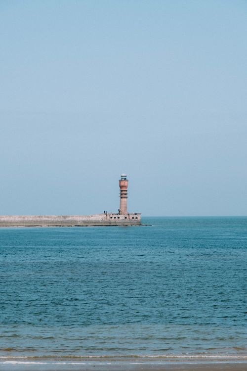 Free A solitary lighthouse stands by a tranquil sea under a clear blue sky, capturing coastal serenity. Stock Photo