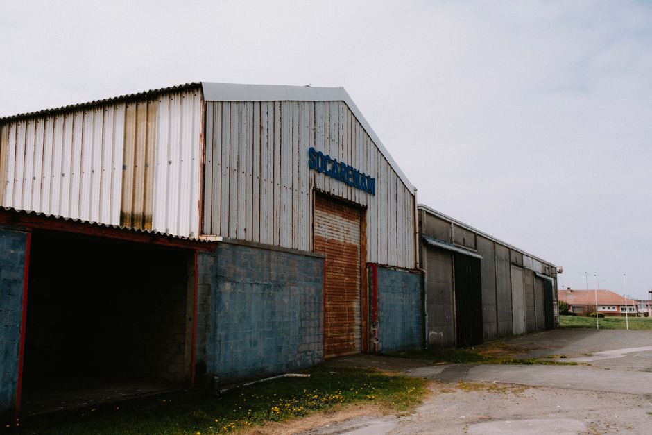 Exterior view of a weathered and rusty industrial warehouse with a roll-up door under a cloudy sky.