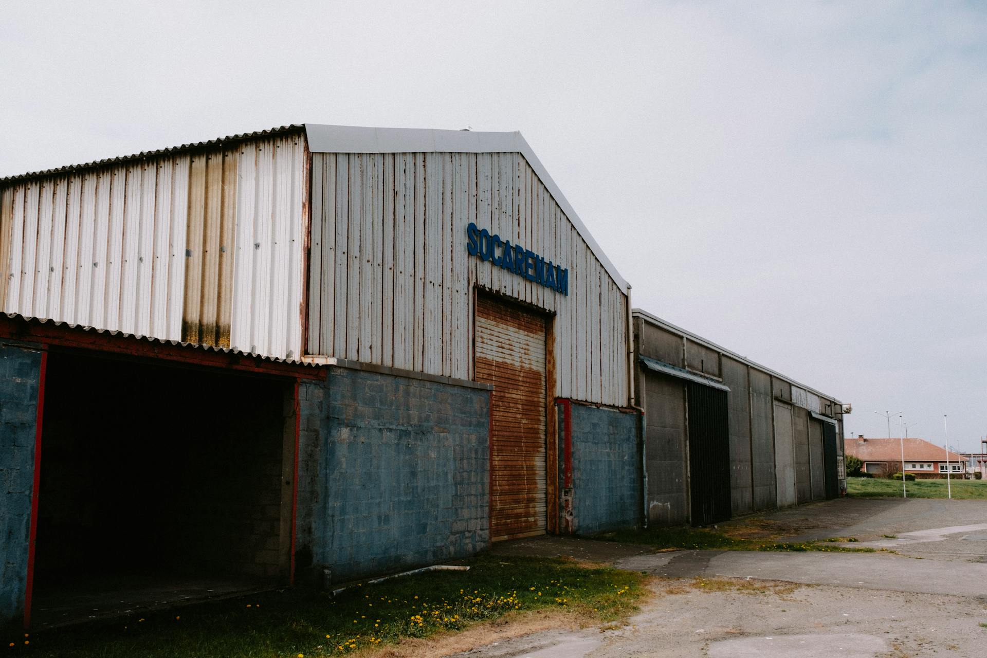 Exterior view of a weathered and rusty industrial warehouse with a roll-up door under a cloudy sky.