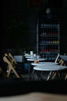 A dimly lit cafe with empty tables and chairs, featuring a solitary iced coffee on a marble table.