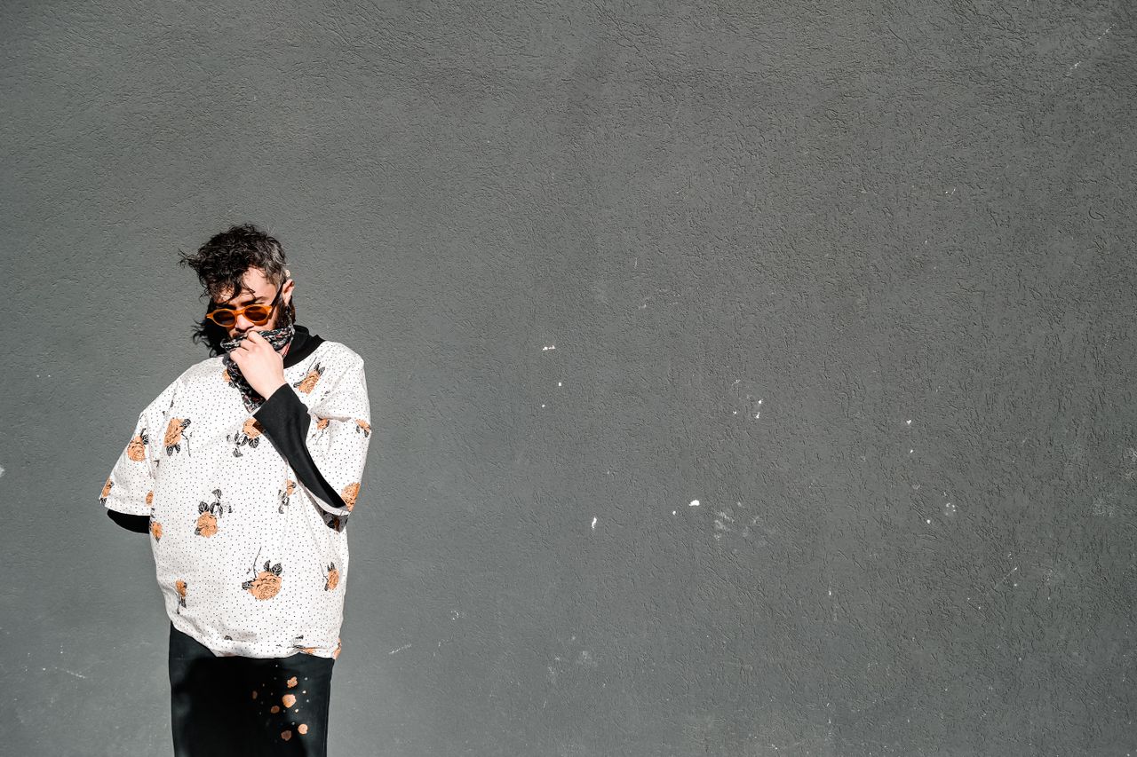 fashion forward man in sunglasses standing against a grey wall wearing