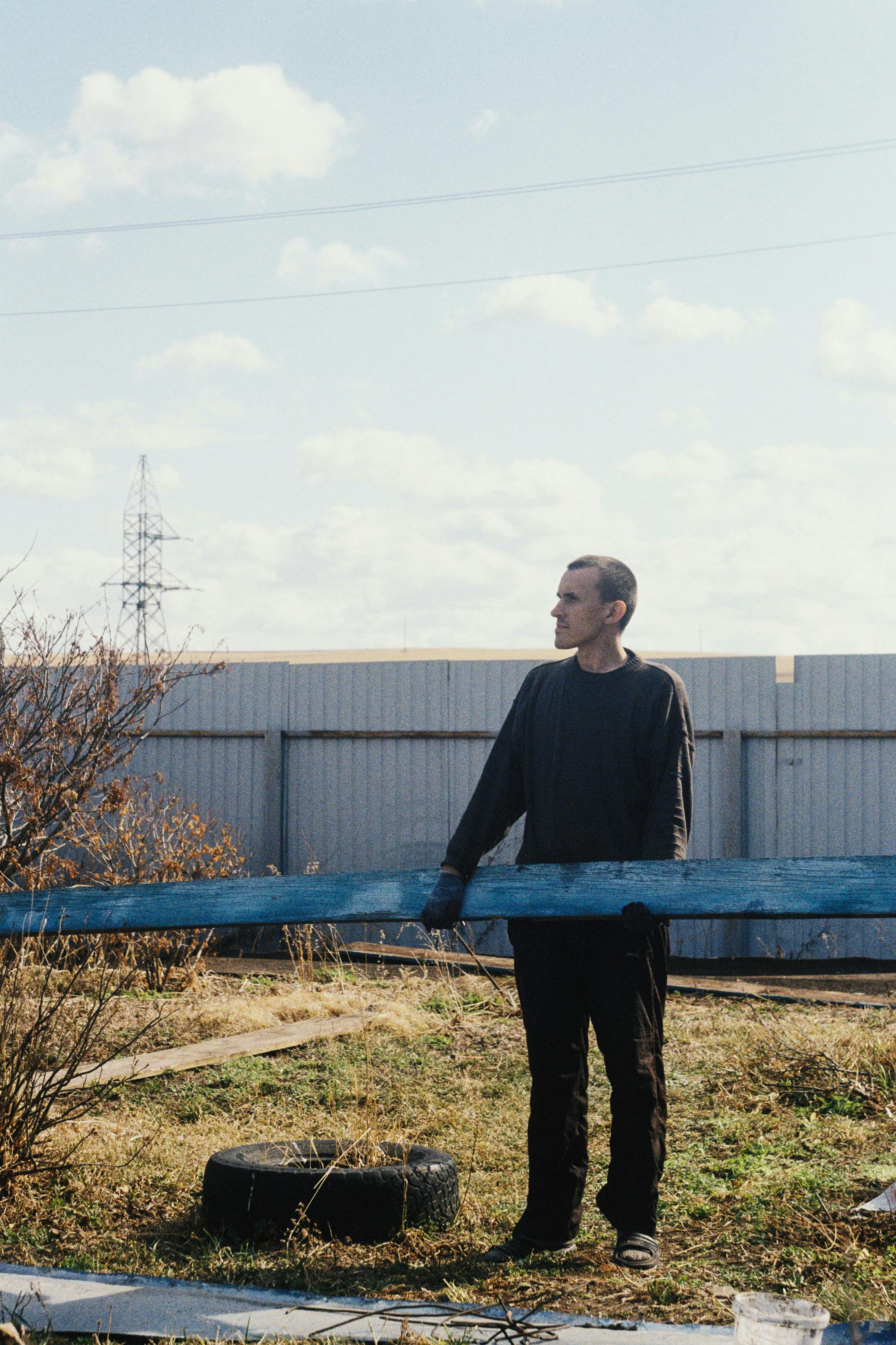 Free A man in black long sleeves stands alone in a grassy, industrial outdoor setting, looking afar. Stock Photo