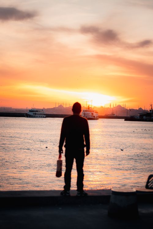 Free A man silhouetted against a vibrant sunset, watching ferries on the water. Stock Photo