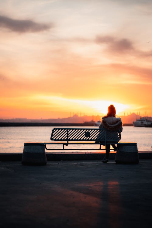 Back View Shot of a Woman Sitting on a Metal Bench while Looking at the Golden Sky