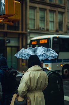 A woman in a raincoat stands under an umbrella at a New York City street corner on a rainy day.