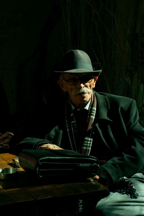 Free Elderly man with glasses and mustache sitting at an outdoor table, classic attire with hat and scarf accentuate a vintage style. Stock Photo
