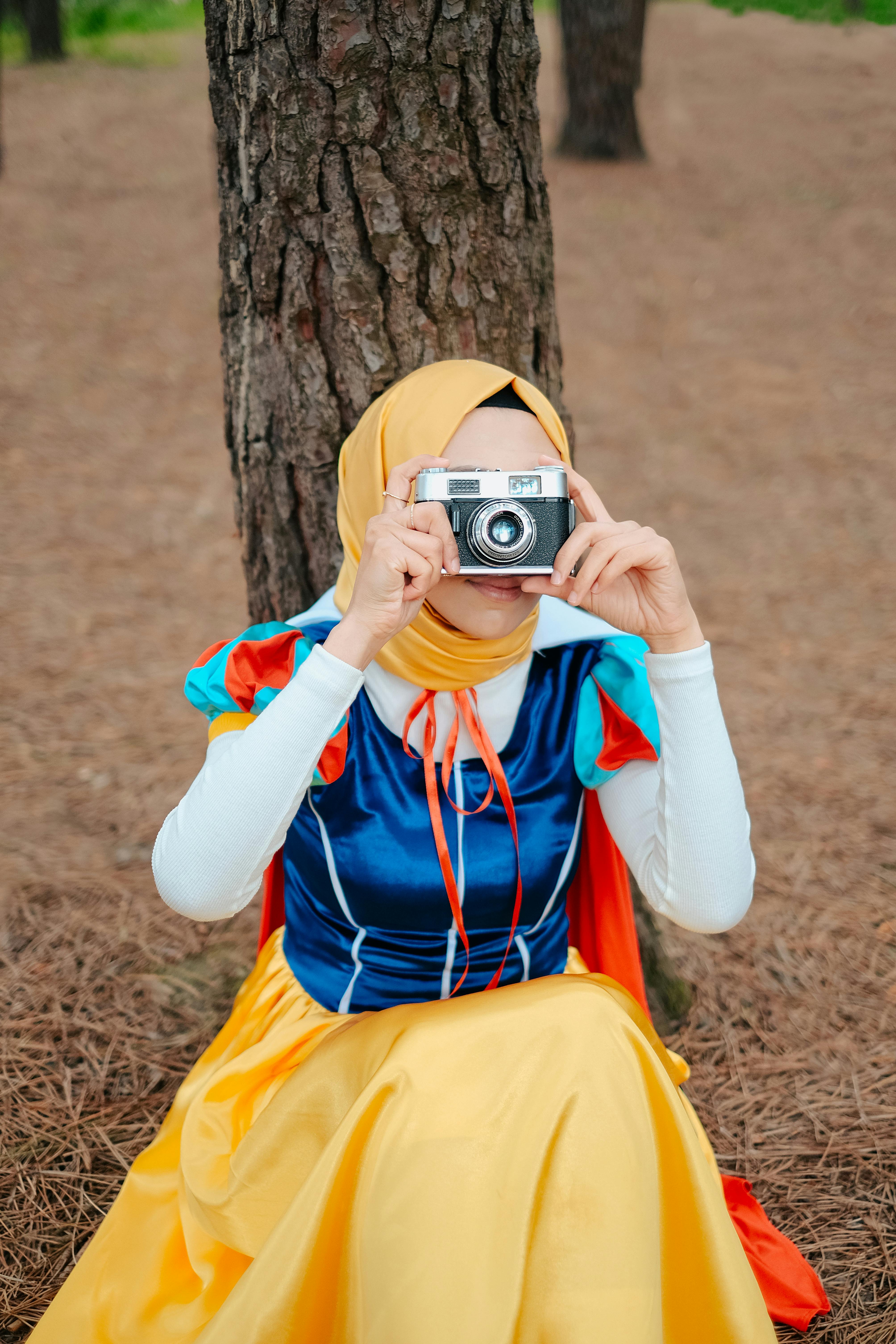 A woman in a Snow White costume sits under a tree taking a picture with an analog camera.