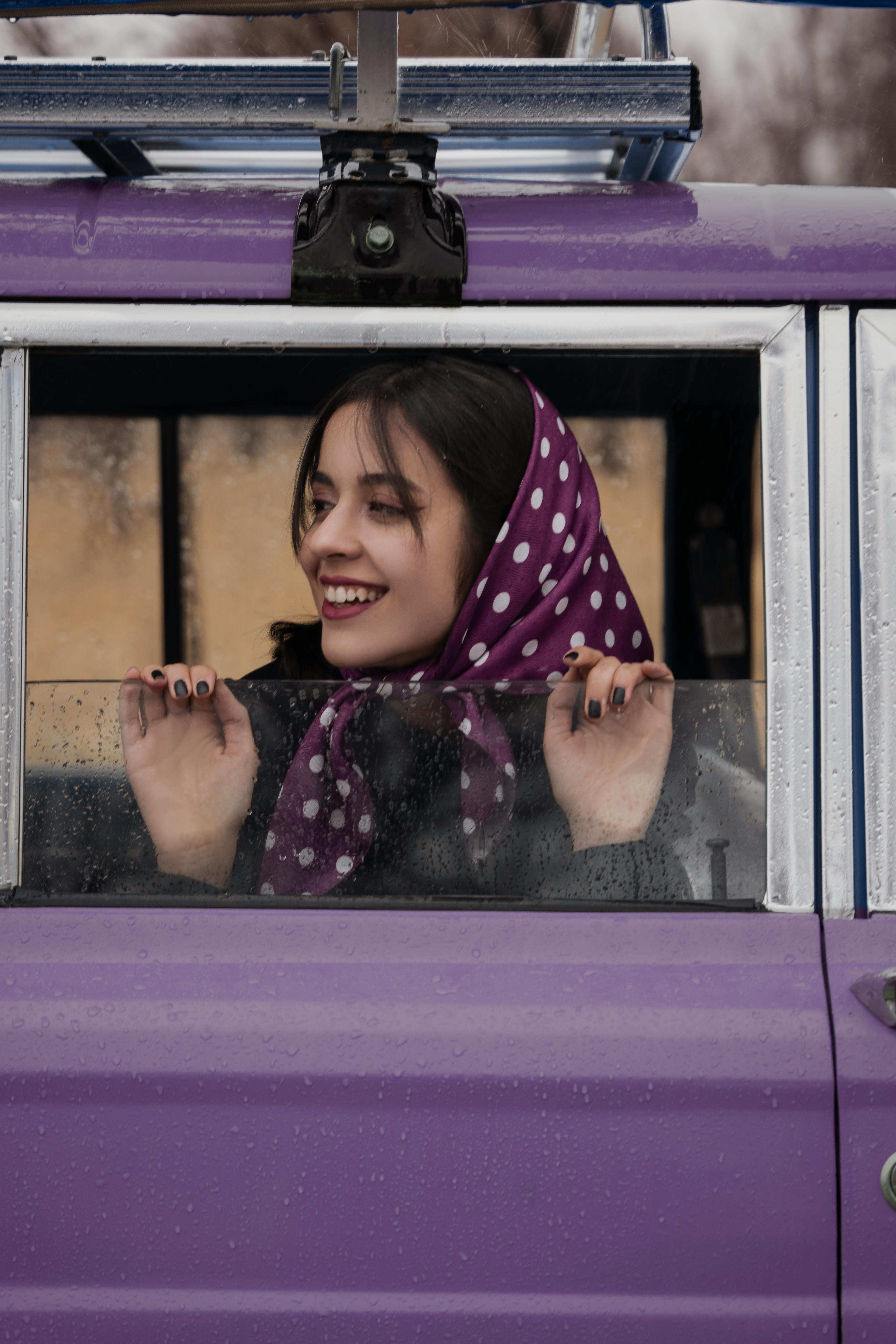 Young woman smiling through a car window, wearing a vibrant polka dot scarf.