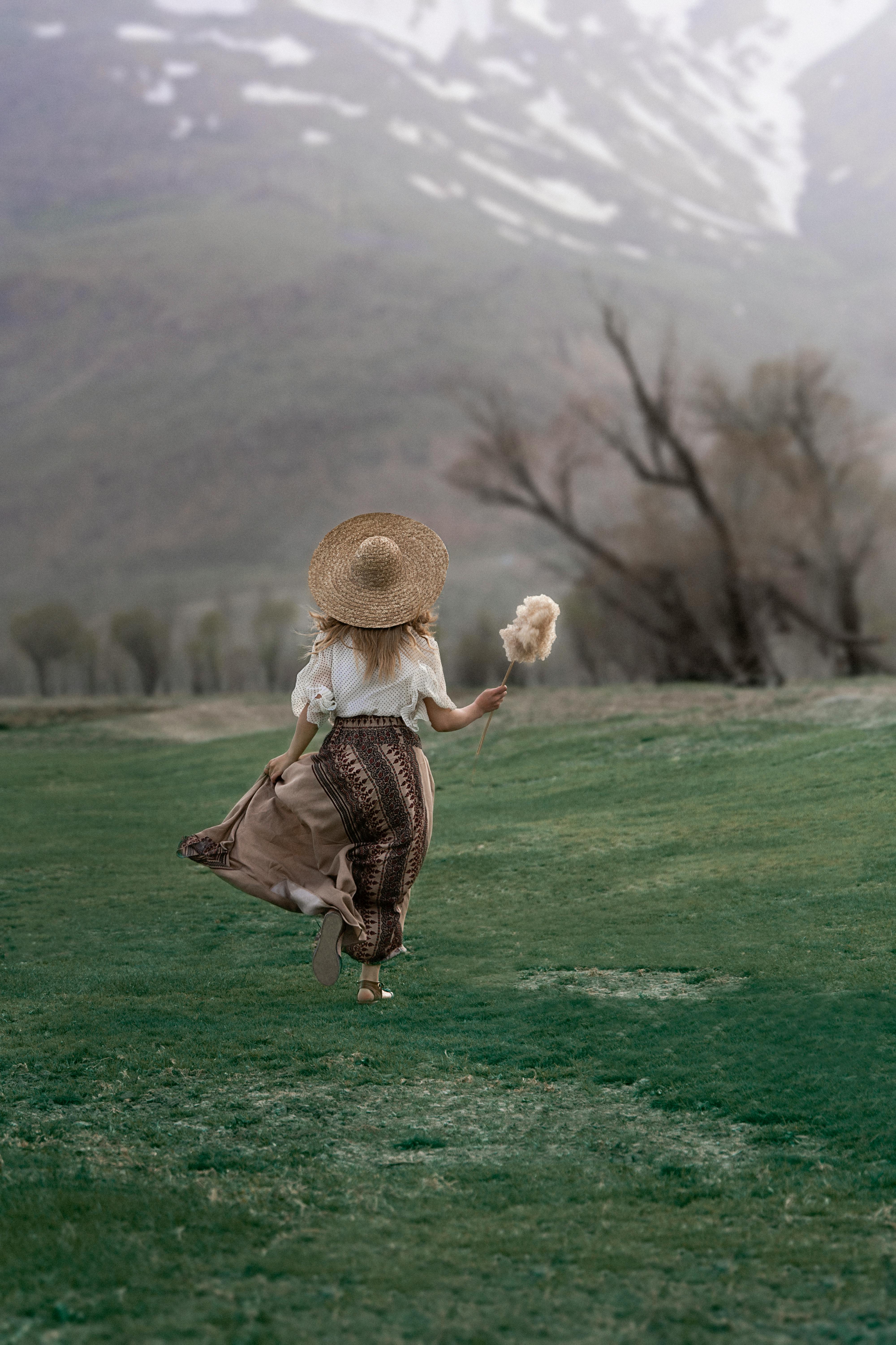 A woman in a straw hat runs through a lush green meadow with mountains in the background.