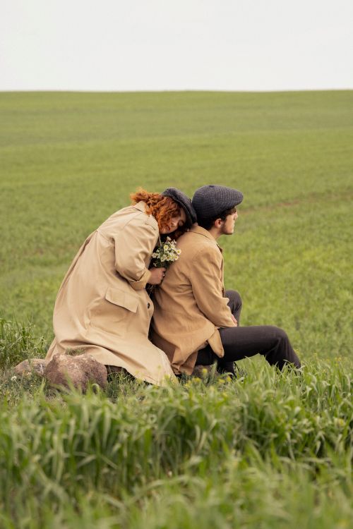 Free Couple sitting together in a lush green field, embracing with coats and hats. Stock Photo