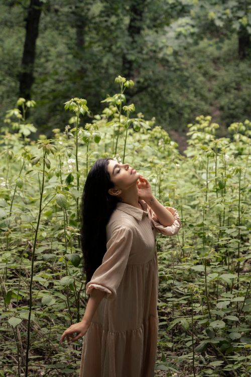 Free A serene woman in a brown dress standing among lush green plants in a forest setting. Stock Photo
