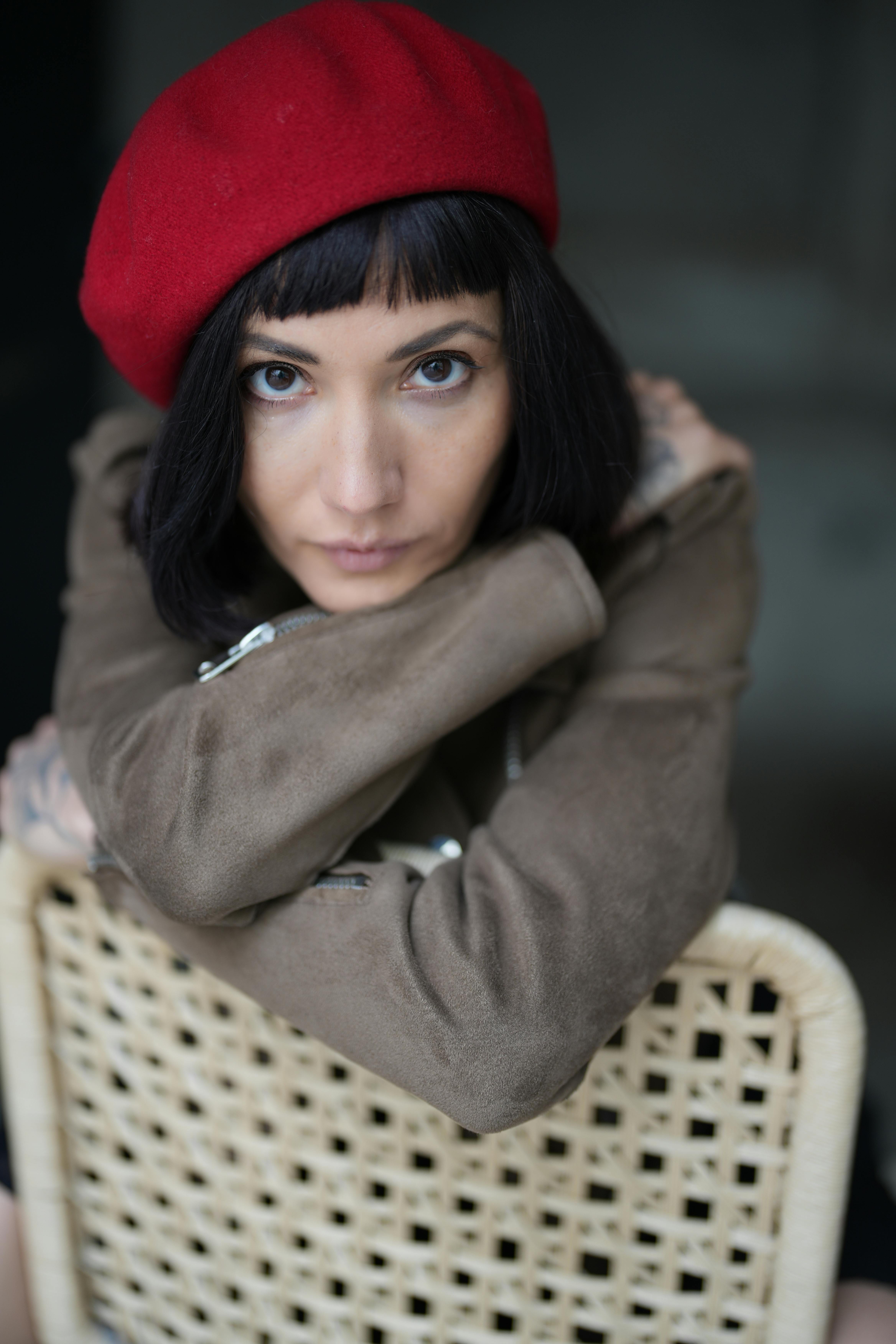 Portrait of a stylish woman in a red beret and brown coat leaning on a woven chair indoors.