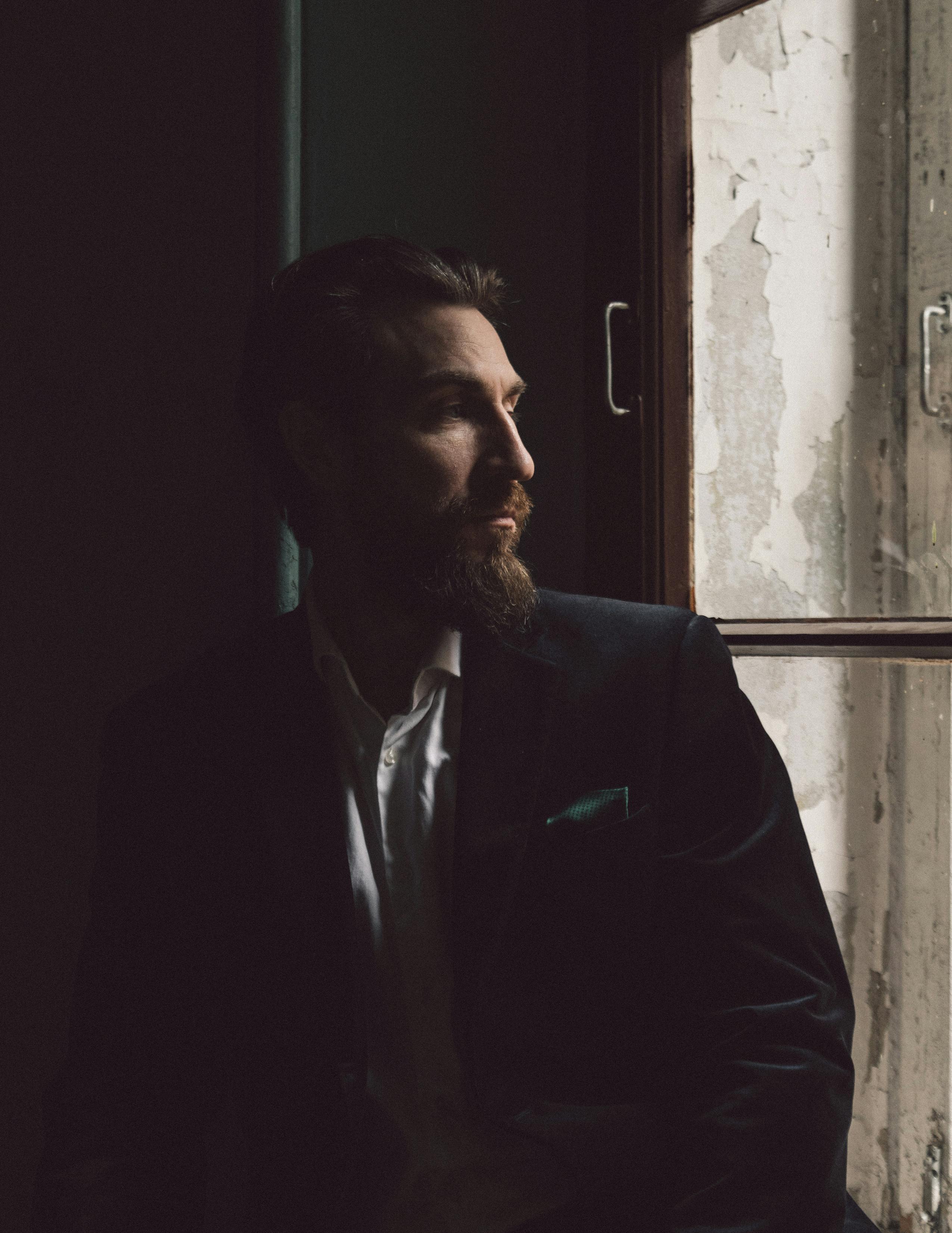 Stylish bearded man in a black suit gazing thoughtfully out a window in low light.