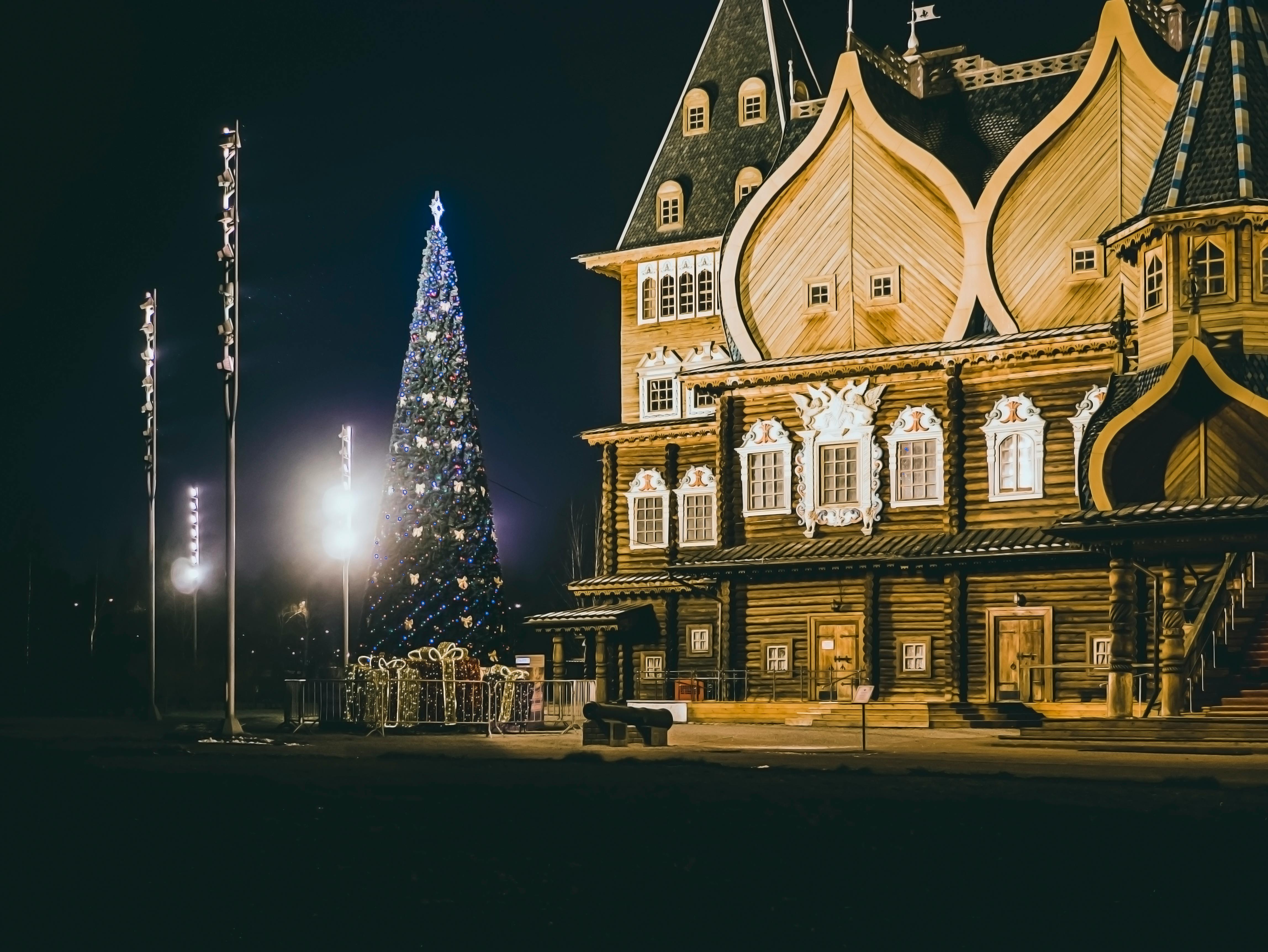 一棵装饰精美的圣诞树旁，矗立着一栋华丽的木制建筑，在夜间灯火通明