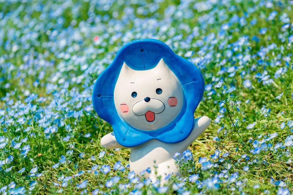 青いトリムのある愛らしいアニメキャラクターが、青い花が咲く明るい草原にいます。