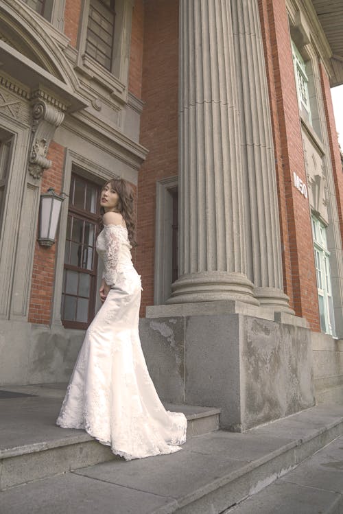 Free Woman in White Wedding Gown Standing Near White Concrete Building Stock Photo