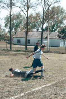 Young men playing a football match on a rural village field with rustic buildings in the background.