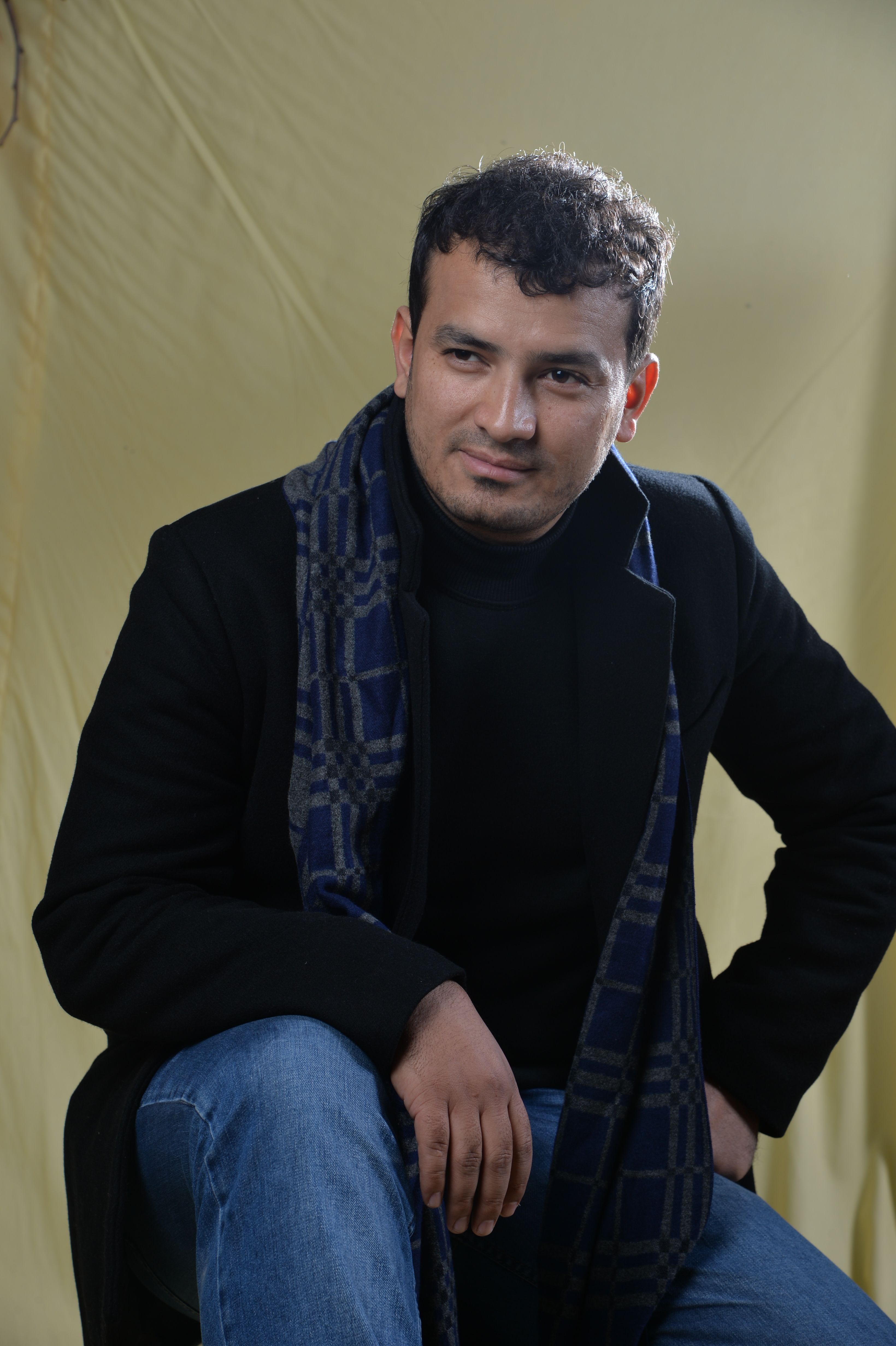 Man in a stylish black blazer and scarf posing casually against a yellow background.